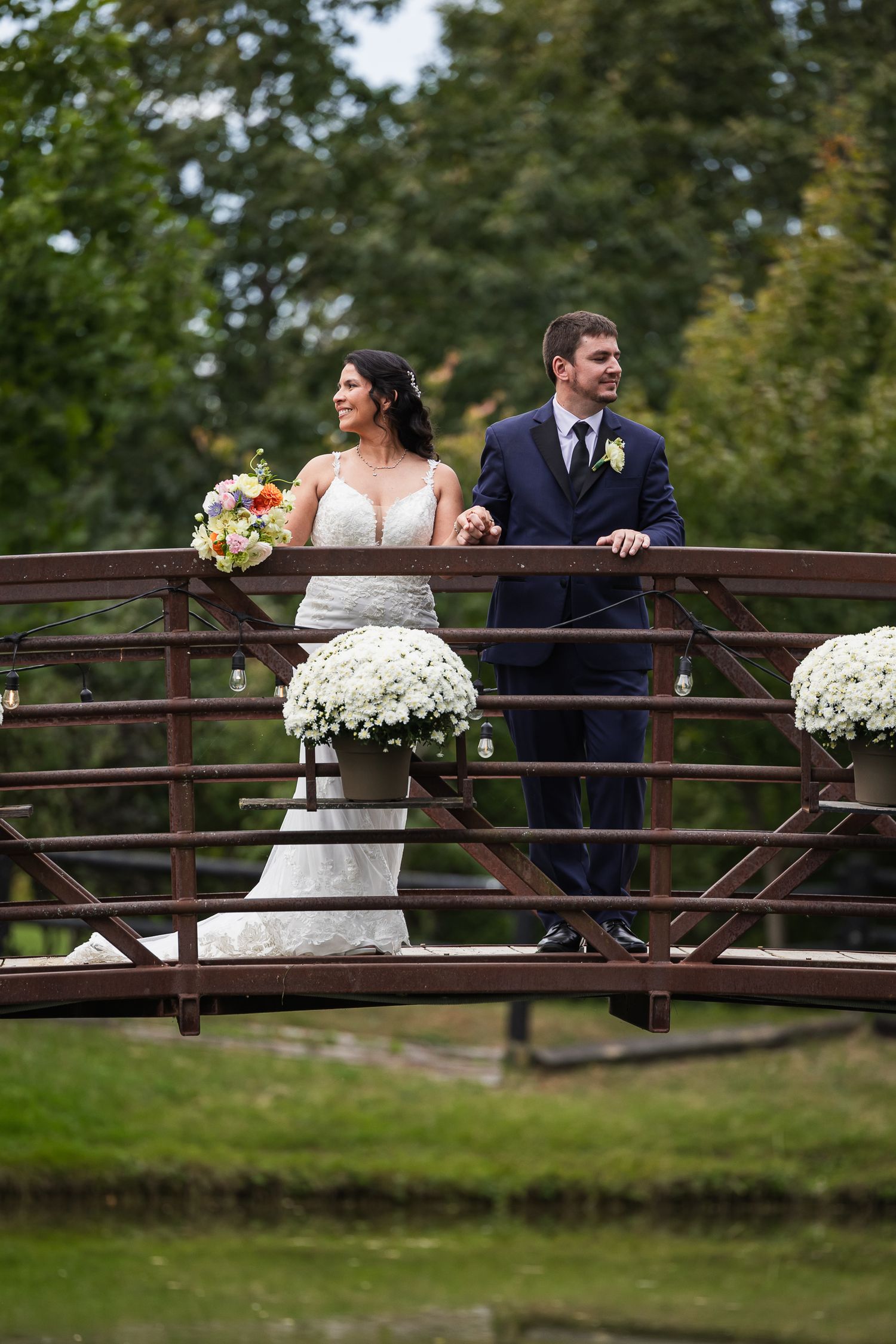 Bride and groom stand on a decorated wooden bridge surrounded by lush greenery during their outdoor wedding.