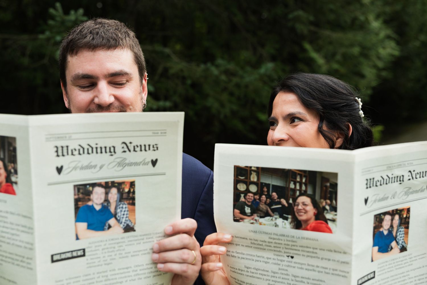 Smiling bride and groom playfully peek over personalized Wedding News newspapers at an outdoor wedding.