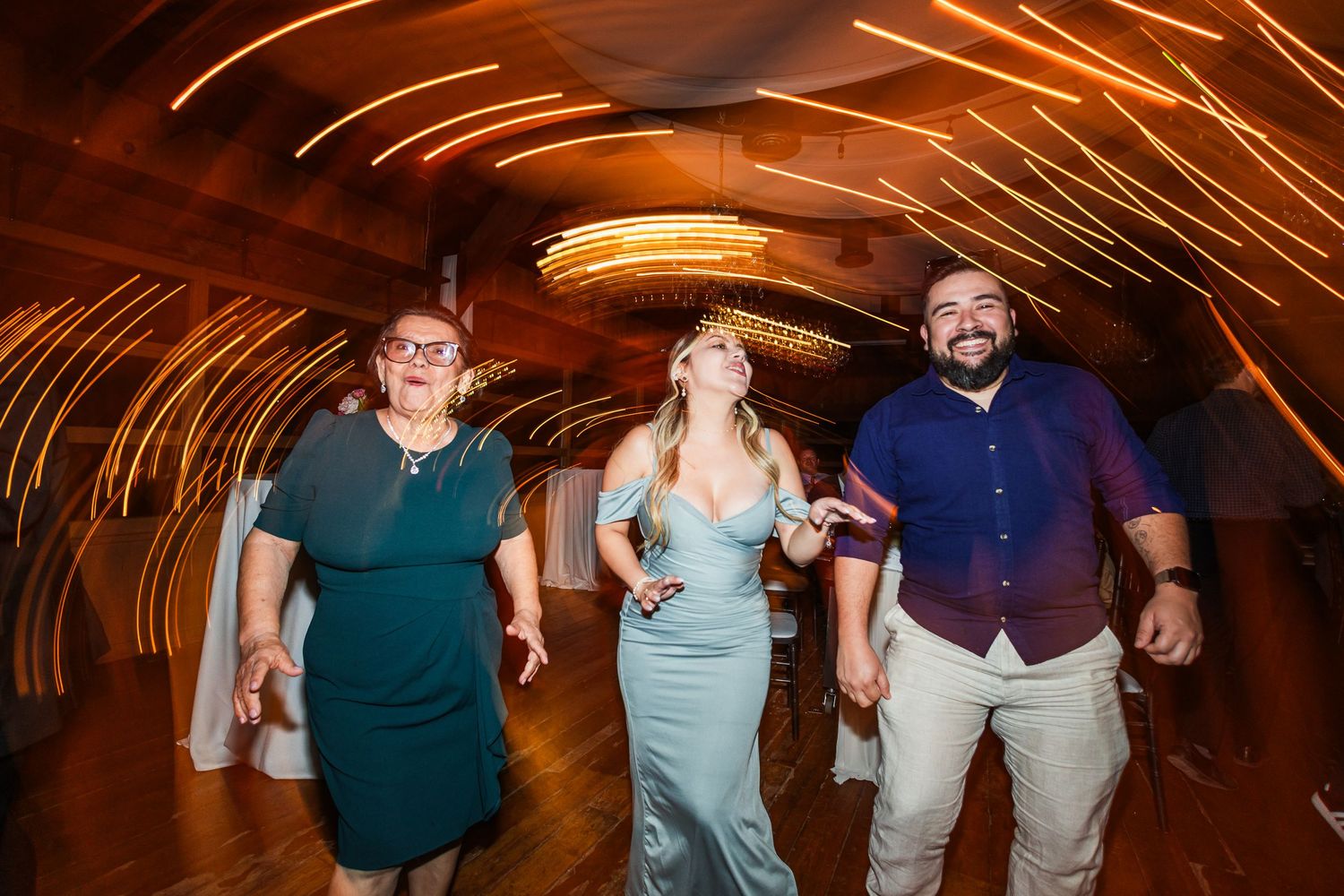 Three guests dance joyfully at a wedding reception with colorful light trails swirling in the background.