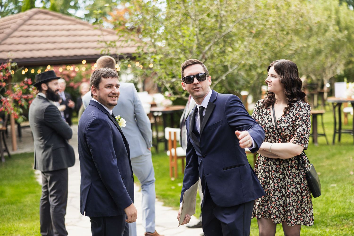 Groomsmen in navy suits and a woman in a floral dress mingle on a sunny outdoor wedding venue lawn.