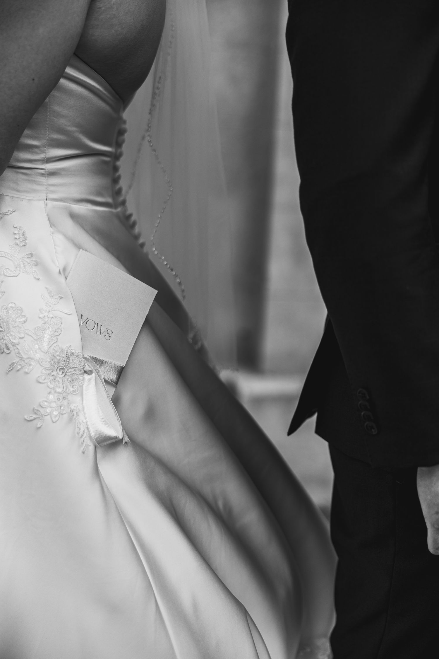 Close-up black and white detail shot of bride's embroidered wedding gown beside groom's dark suit.