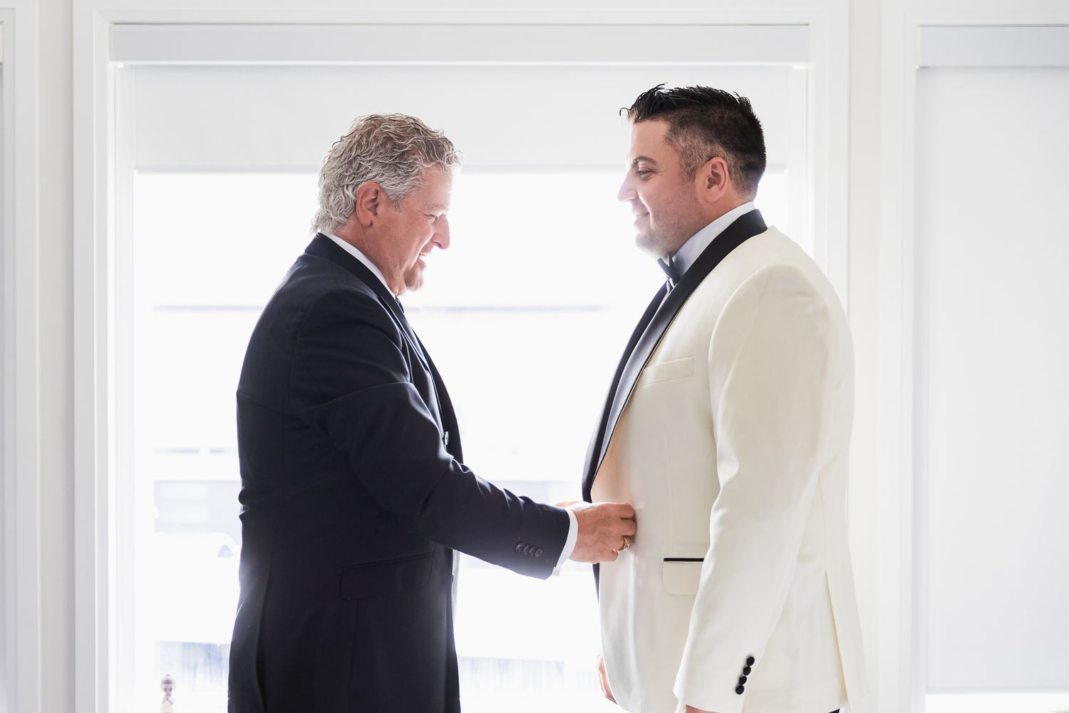 Older man adjusting groom's white tuxedo jacket before wedding ceremony.