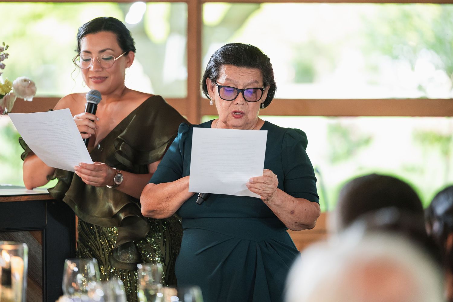 Two women in elegant green dresses deliver heartfelt speeches at an outdoor wedding reception celebration.