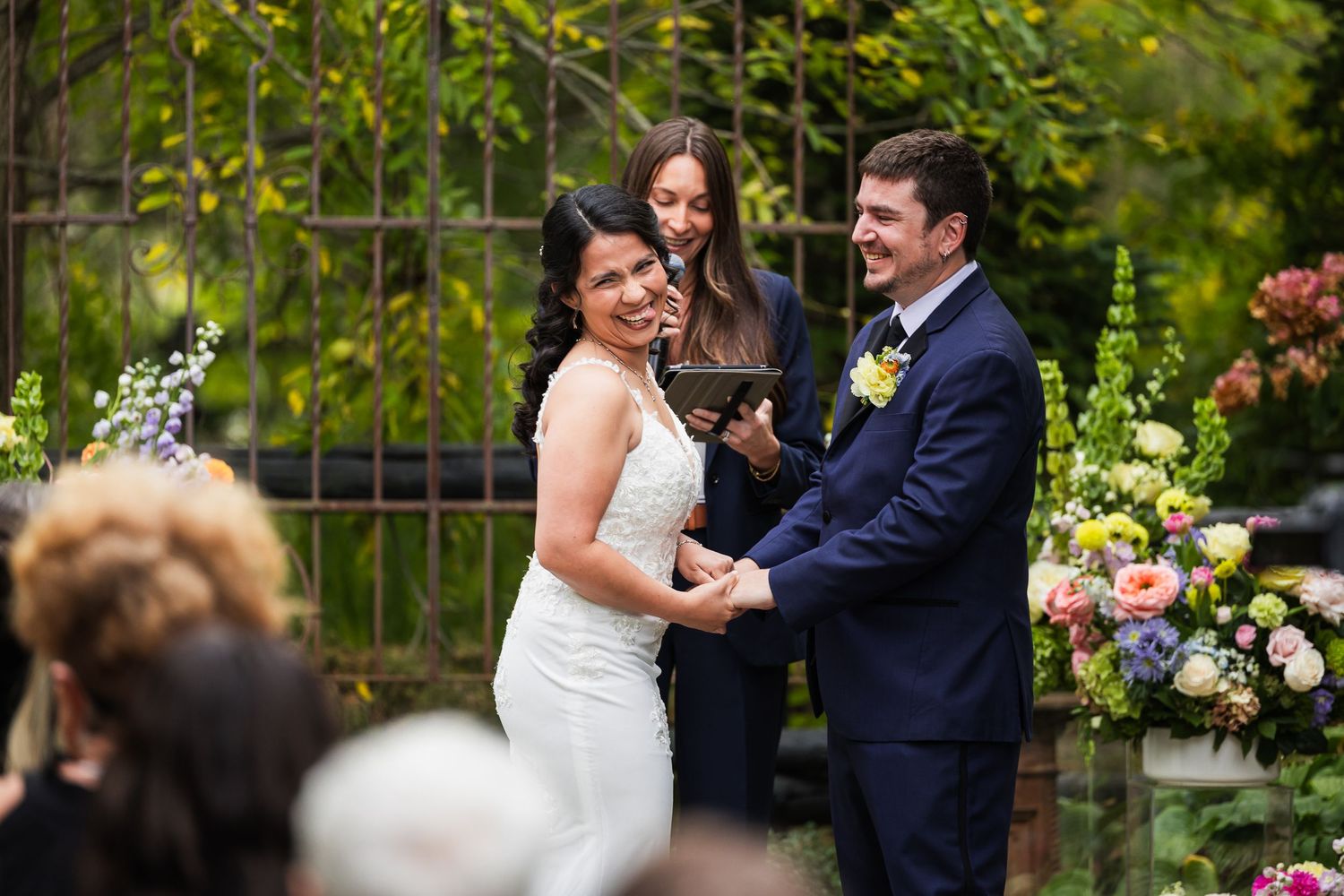 Bride and groom laughing joyfully during outdoor wedding ceremony with officiant and floral arrangements.