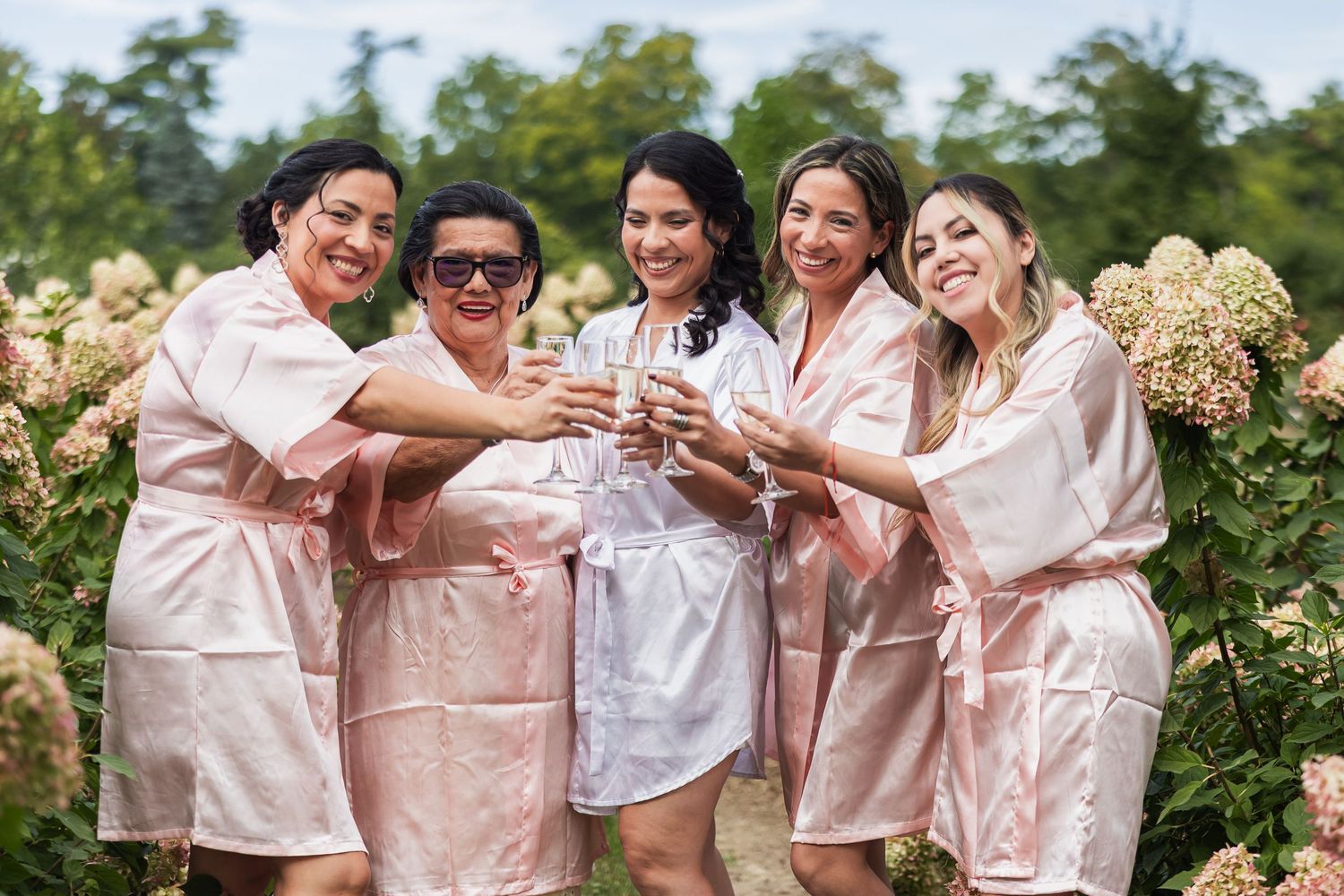 Bridesmaids in pink satin robes toasting champagne with bride in white robe outdoors among blooming hydrangeas.