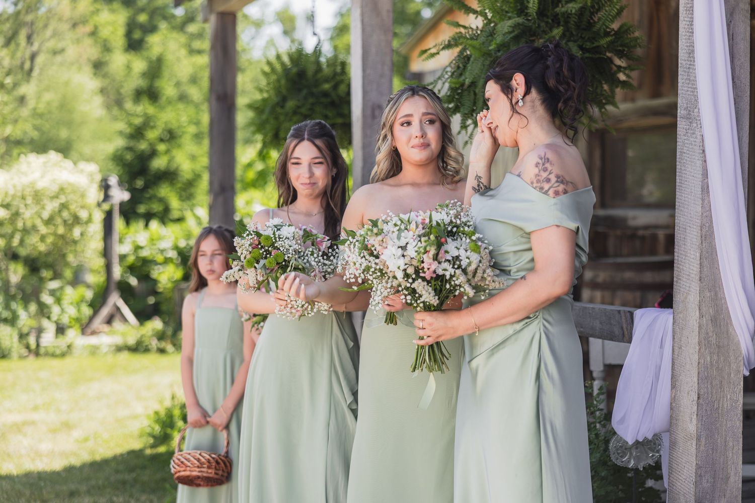 Bridesmaids in sage green off-shoulder gowns hold floral bouquets with flower girl at rustic barn wedding.