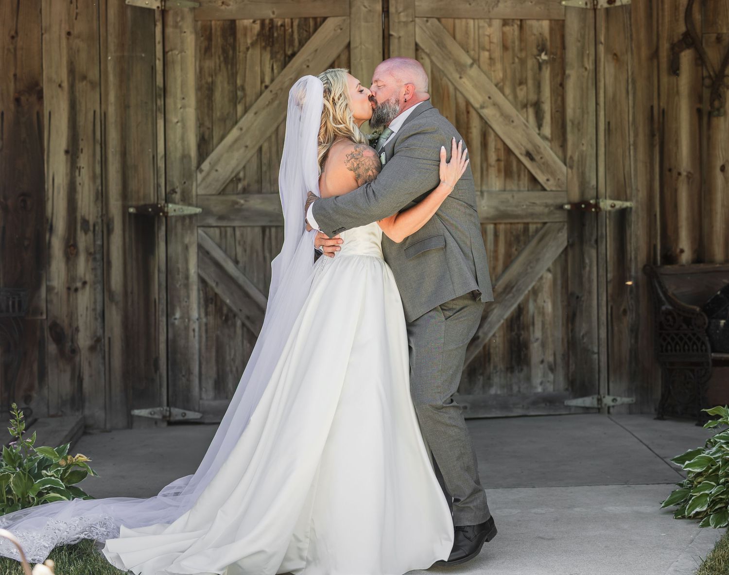 Bride and groom share first kiss in front of rustic barn doors at outdoor wedding ceremony.