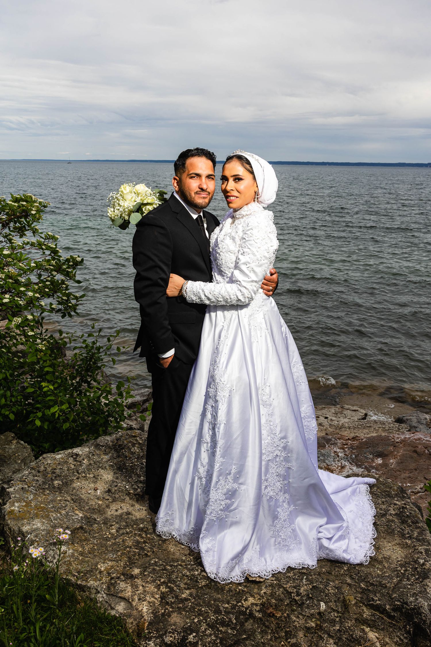 Newlyweds embrace on rocky shoreline with ocean and cloudy sky in background.