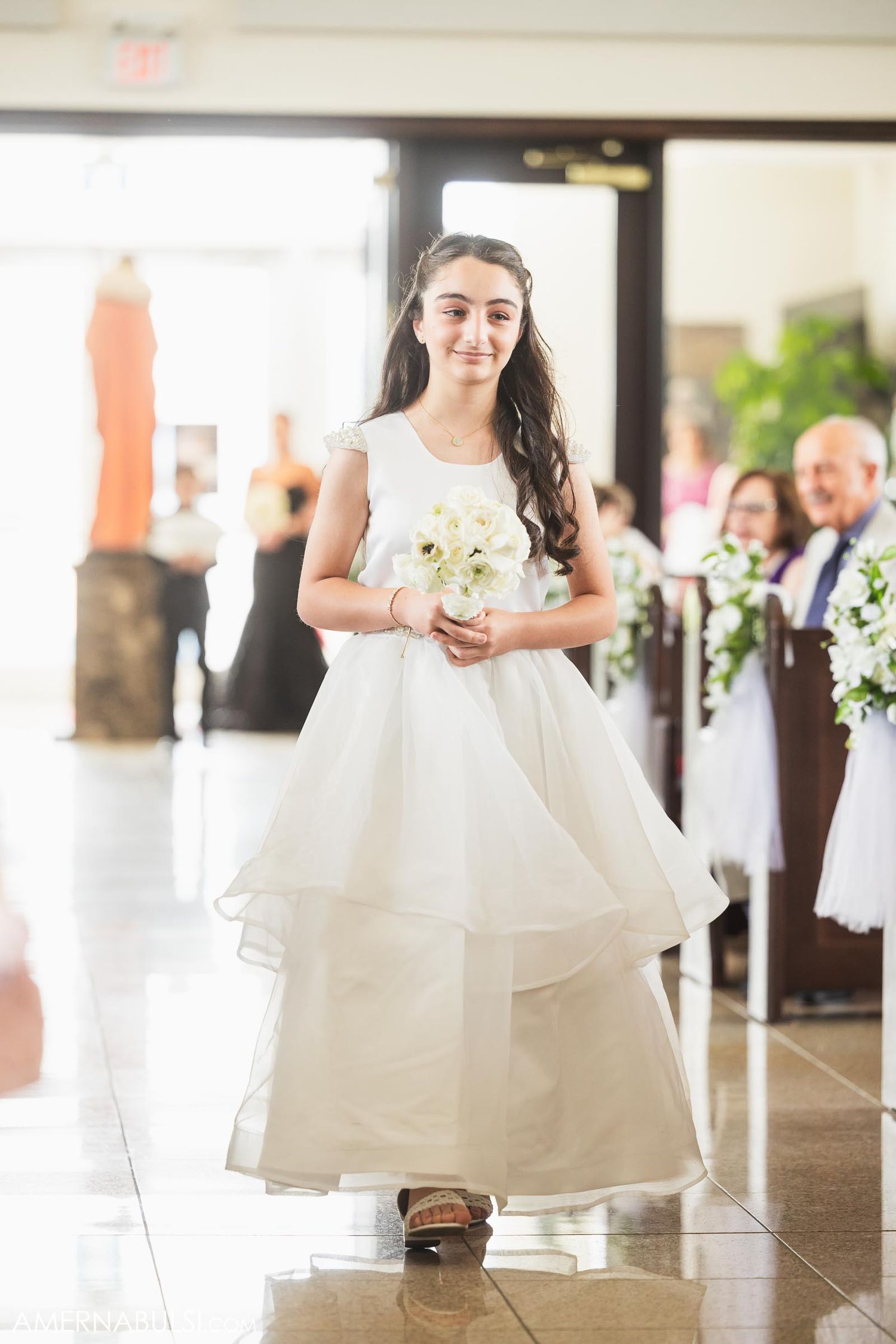 Young flower girl in white layered dress walking down aisle with bouquet.
