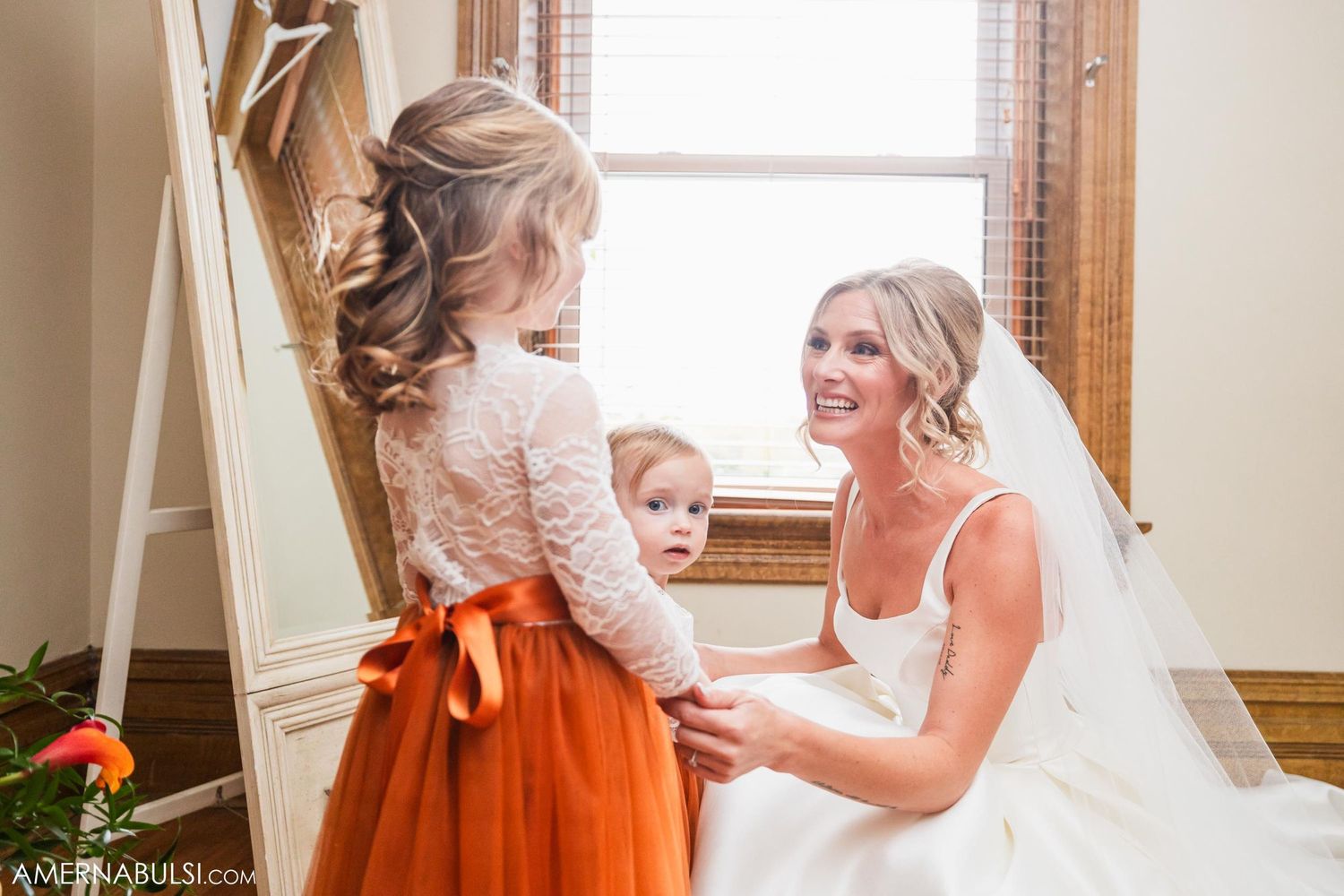 Bride in white wedding dress shares tender moment with young flower girl wearing orange dress and bow.