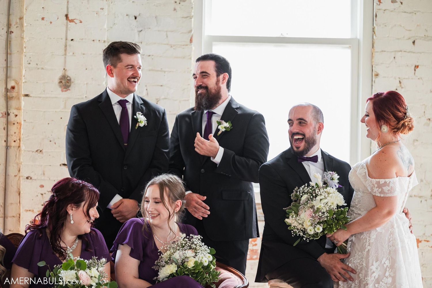 Wedding party laughing together during ceremony in rustic brick venue with bridesmaids in purple dresses.