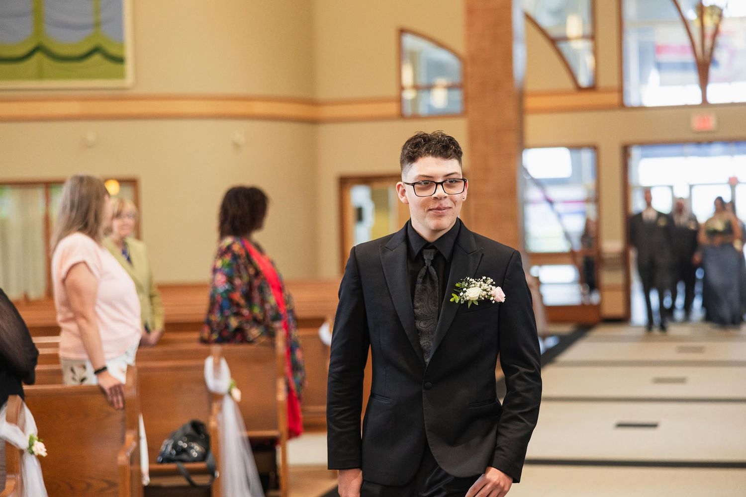A person in a black suit and boutonniere stands in a church during a wedding ceremony.