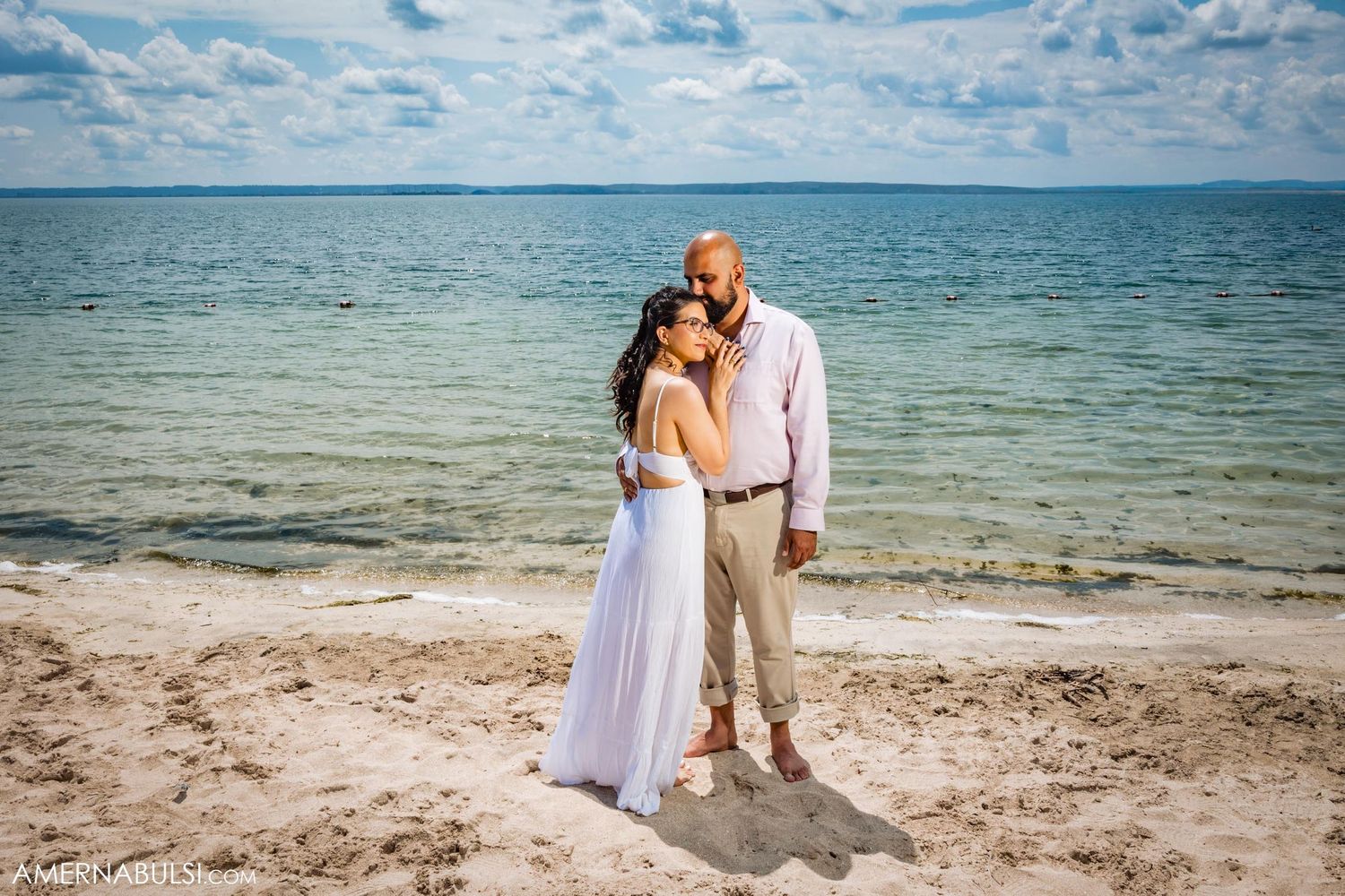 A couple embraces on a beach with blue ocean waters and cloudy sky in the background.