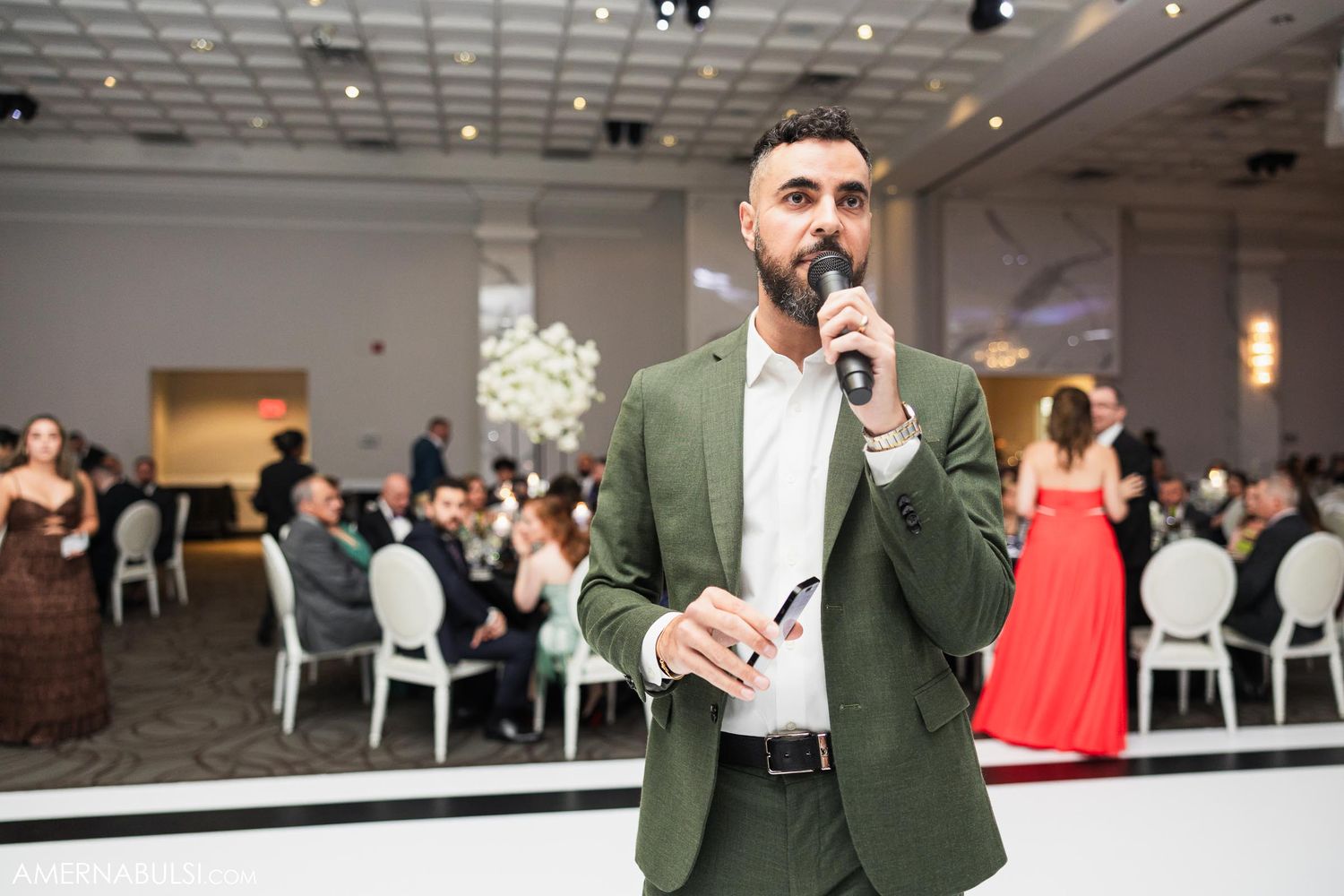 Speaker in green blazer addresses wedding reception guests in modern event space.