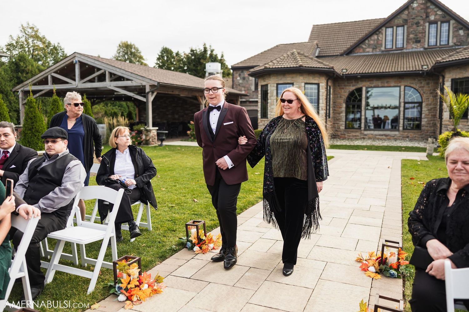 Wedding ceremony processional on stone pathway with autumn decorations and seated guests.