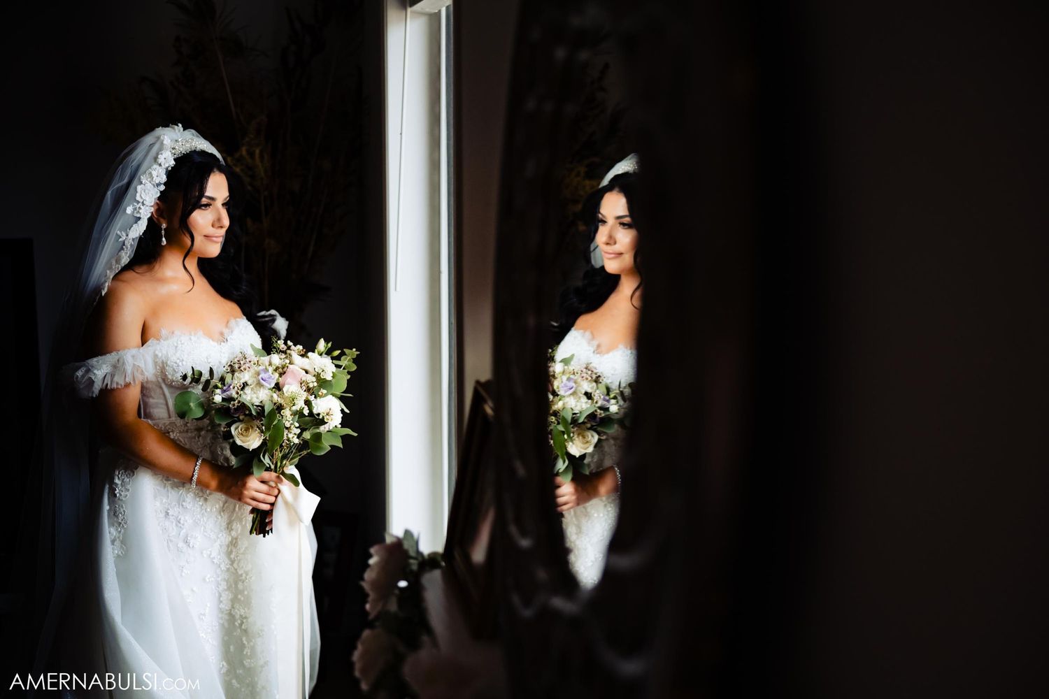 Bride in white off-shoulder gown poses with bouquet next to mirror reflection.