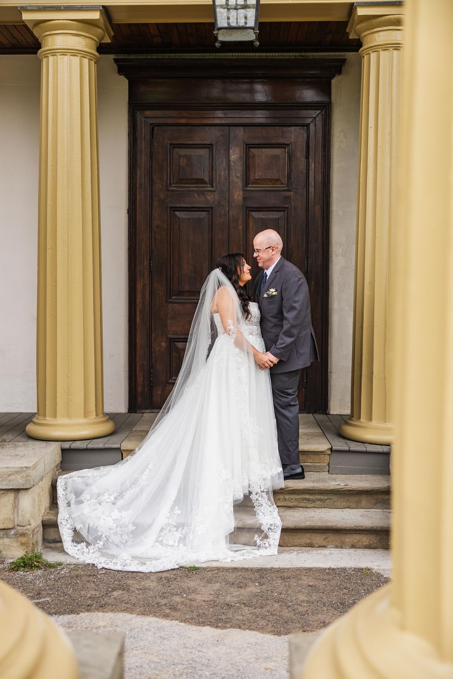 Wedding couple poses in doorway framed by yellow columns at historic mansion entrance.