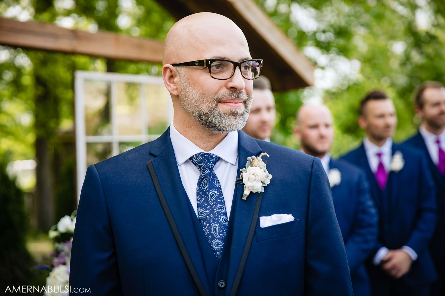 Groomsmen in matching navy blue suits and floral boutonnieres gather outdoors for a wedding photo.