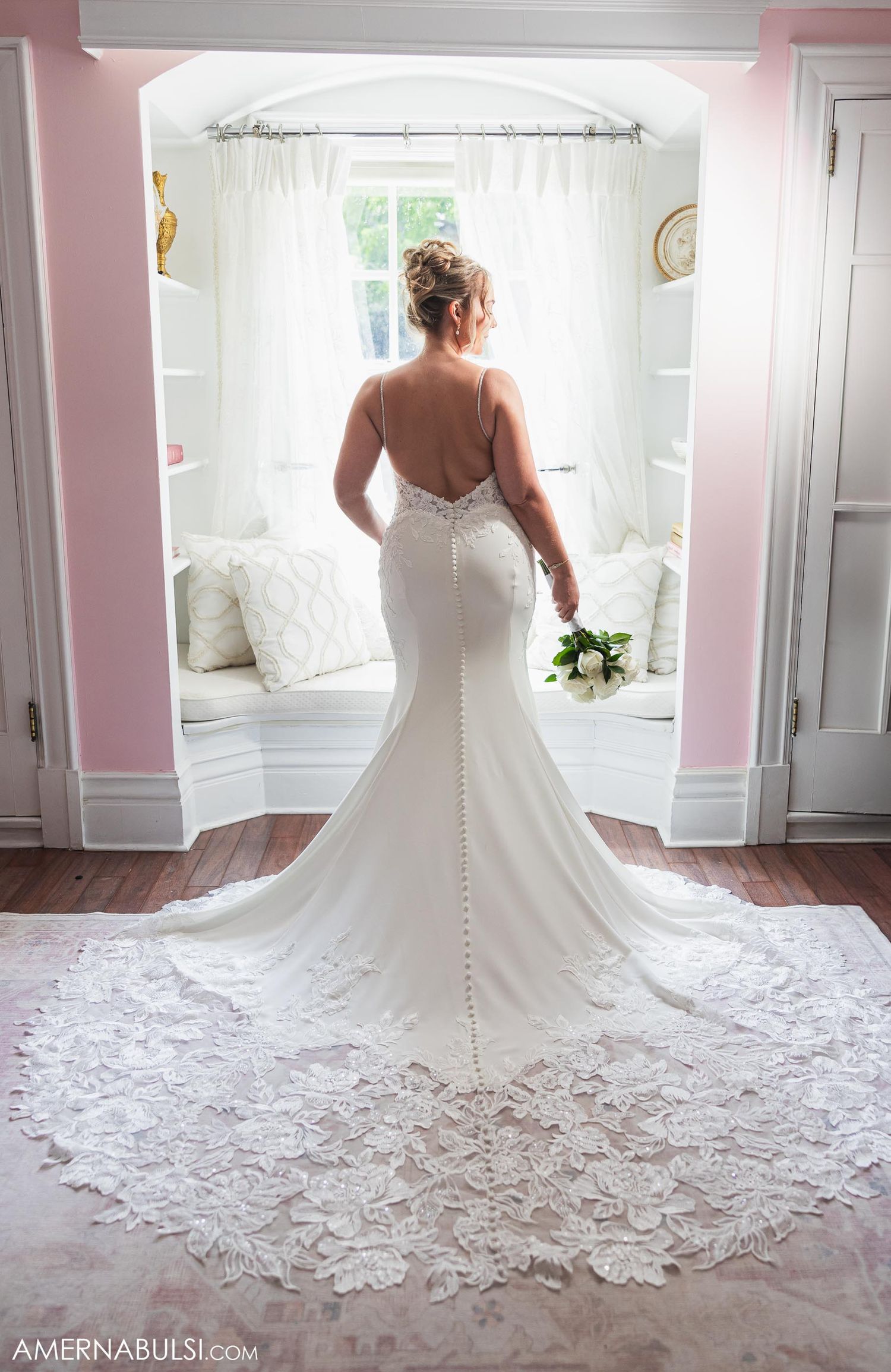 Back view of fitted wedding dress with detailed lace train in pink-walled room.