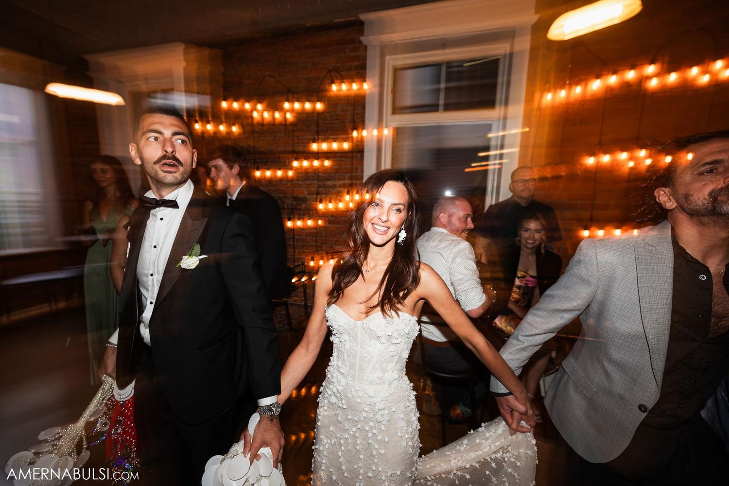 Guests dancing and celebrating at a wedding reception decorated with warm string lights.