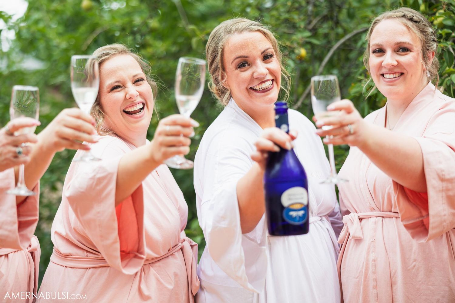 Bridesmaids in pink robes toasting with champagne glasses in an outdoor setting.
