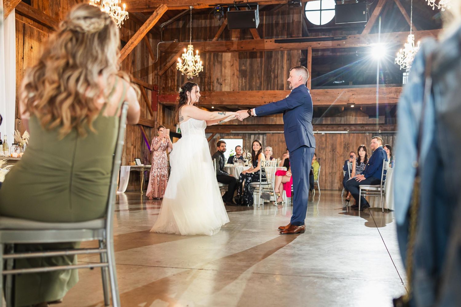 Couple performs their first dance in a rustic barn setting with chandeliers and wooden beams.