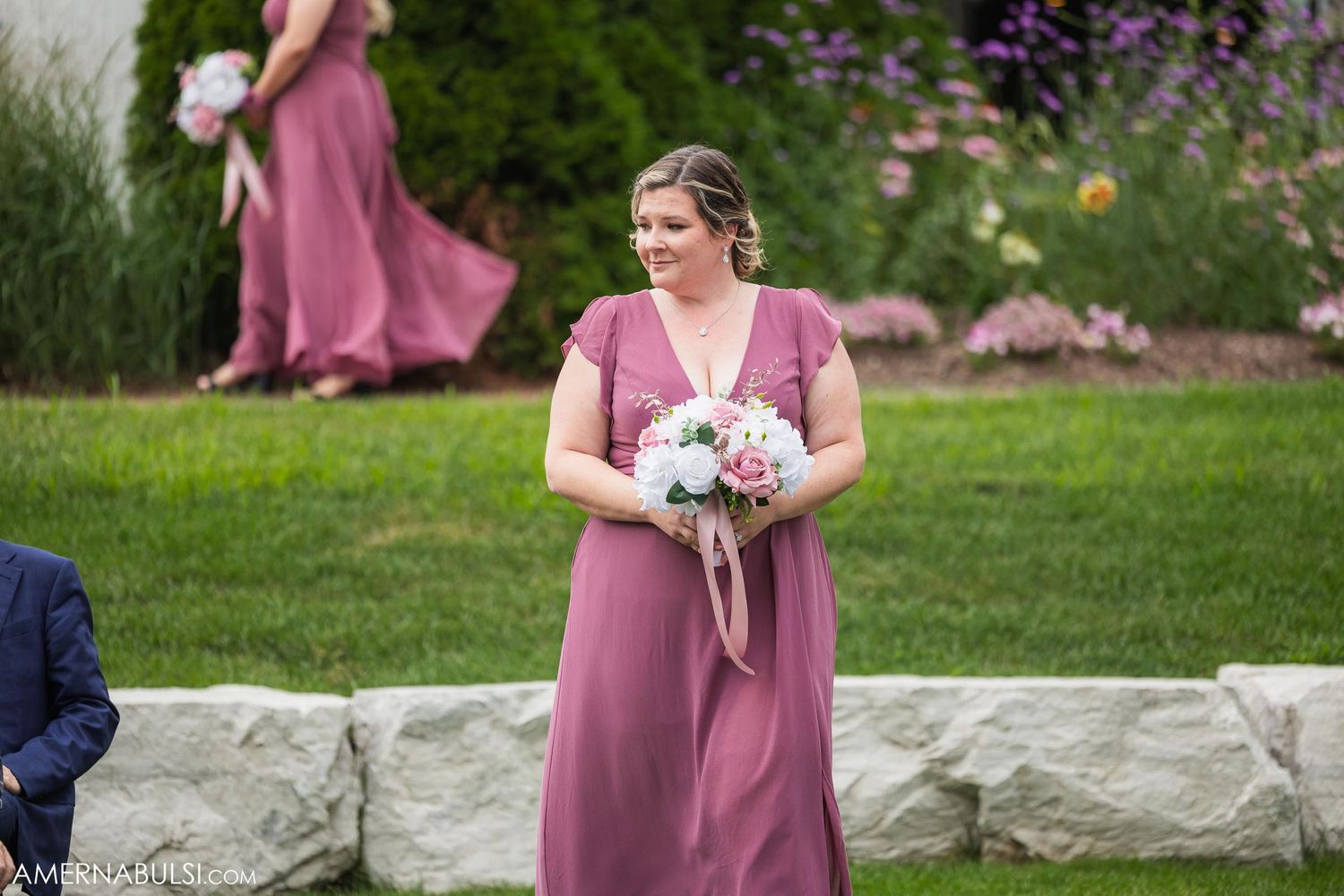 Bridesmaid in mauve dress holds bouquet during outdoor garden wedding ceremony.