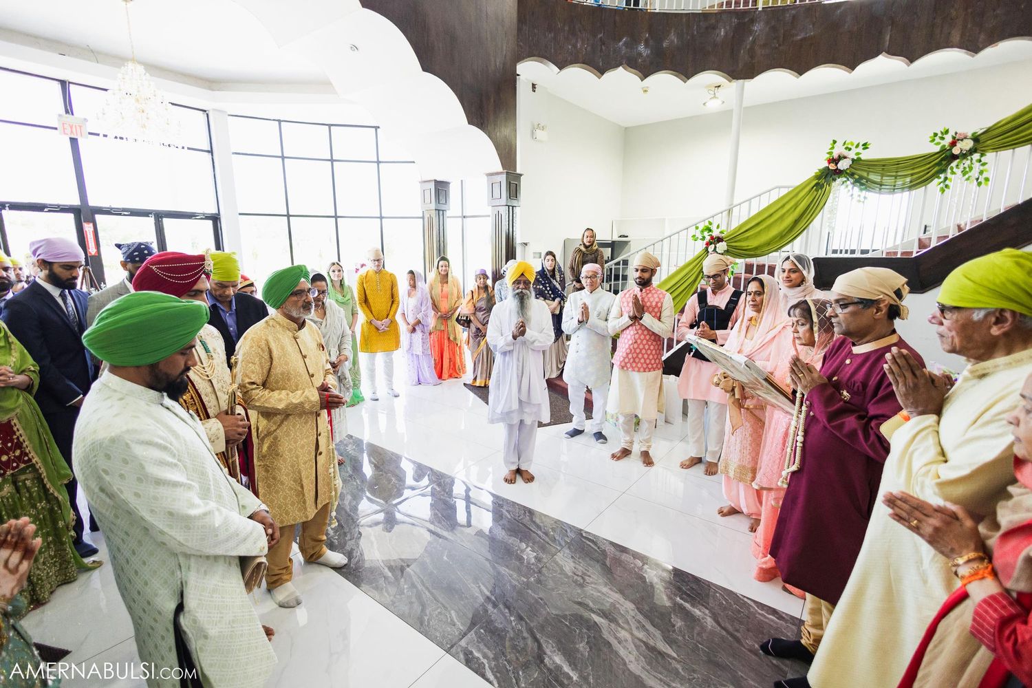 Guests in colorful traditional attire gather inside a bright modern venue for a Sikh wedding celebration.