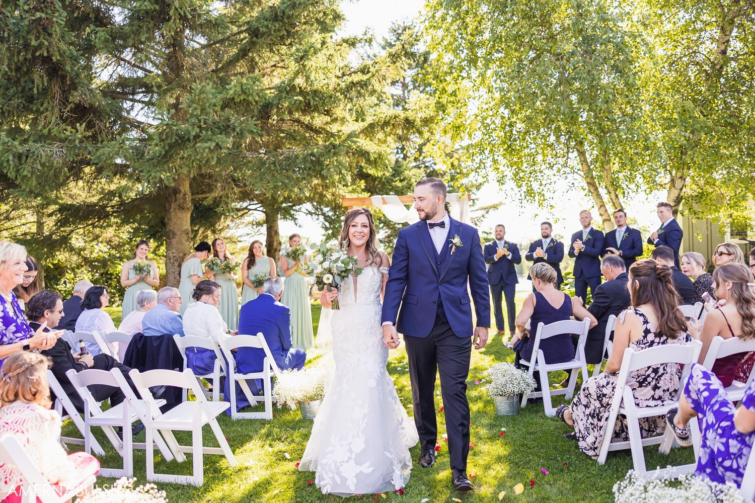 Couple walks down the aisle at sunny outdoor wedding with guests seated on white chairs.