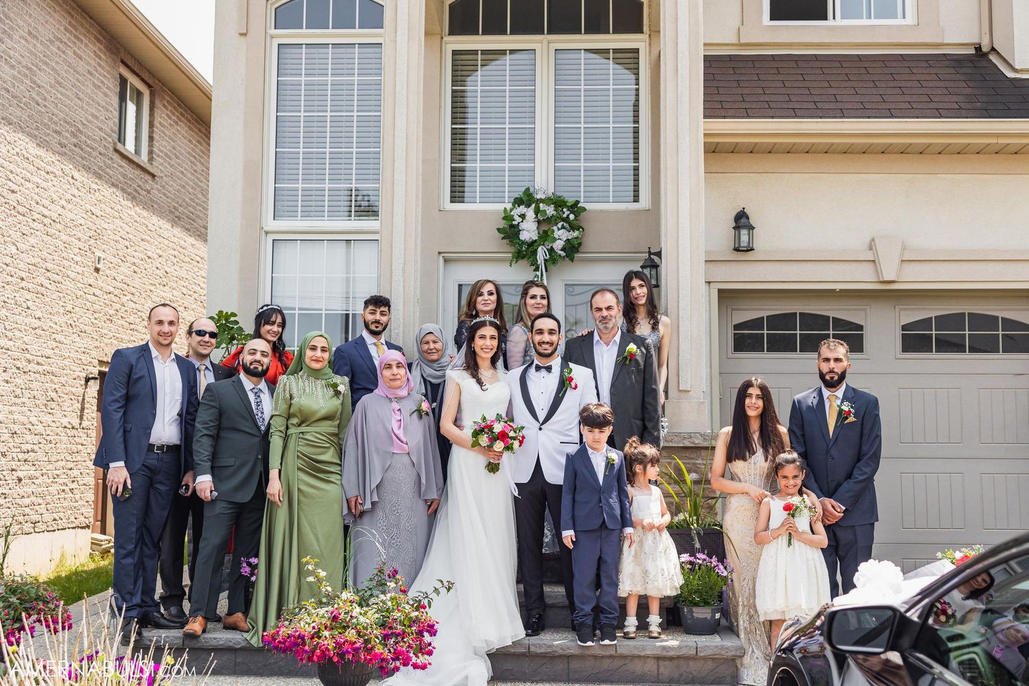 A wedding party poses for a formal group photo outside a large house.