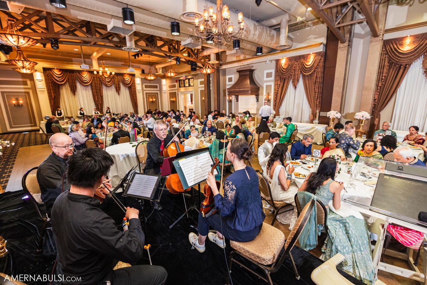 Live orchestra performs at an elegant wedding reception in a grand ballroom with chandeliers and draped decor.
