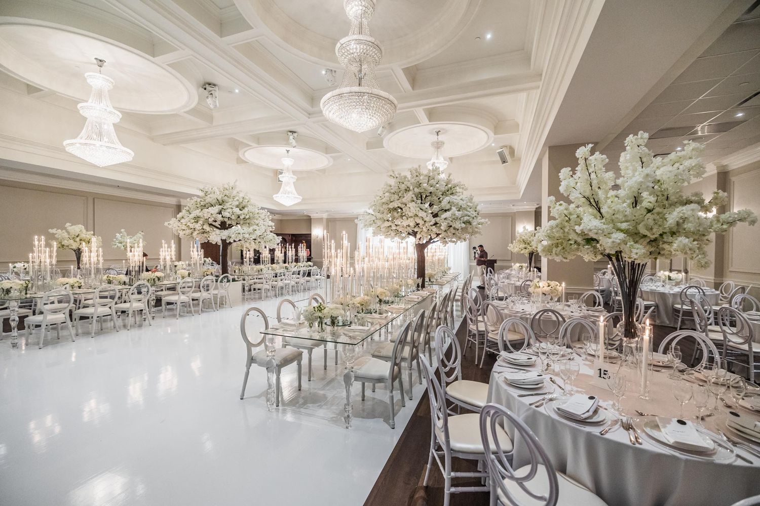 Luxurious wedding reception hall with crystal chandeliers and tall white floral centerpieces.