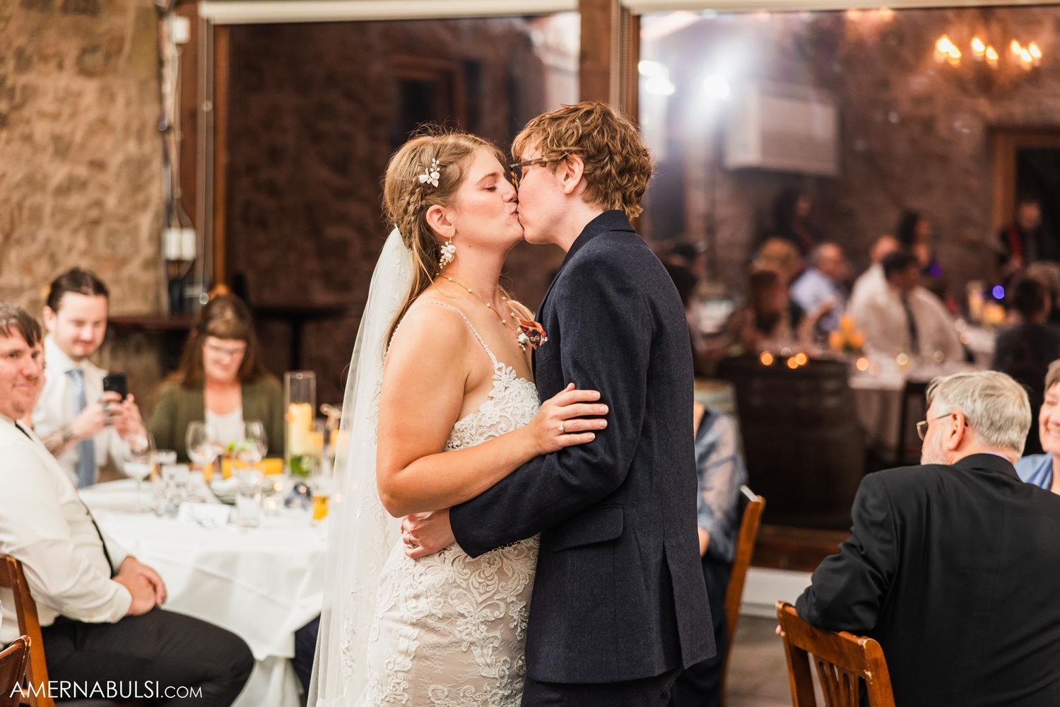 A couple shares their first kiss during a wedding reception in a rustic stone venue with guests seated at tables.