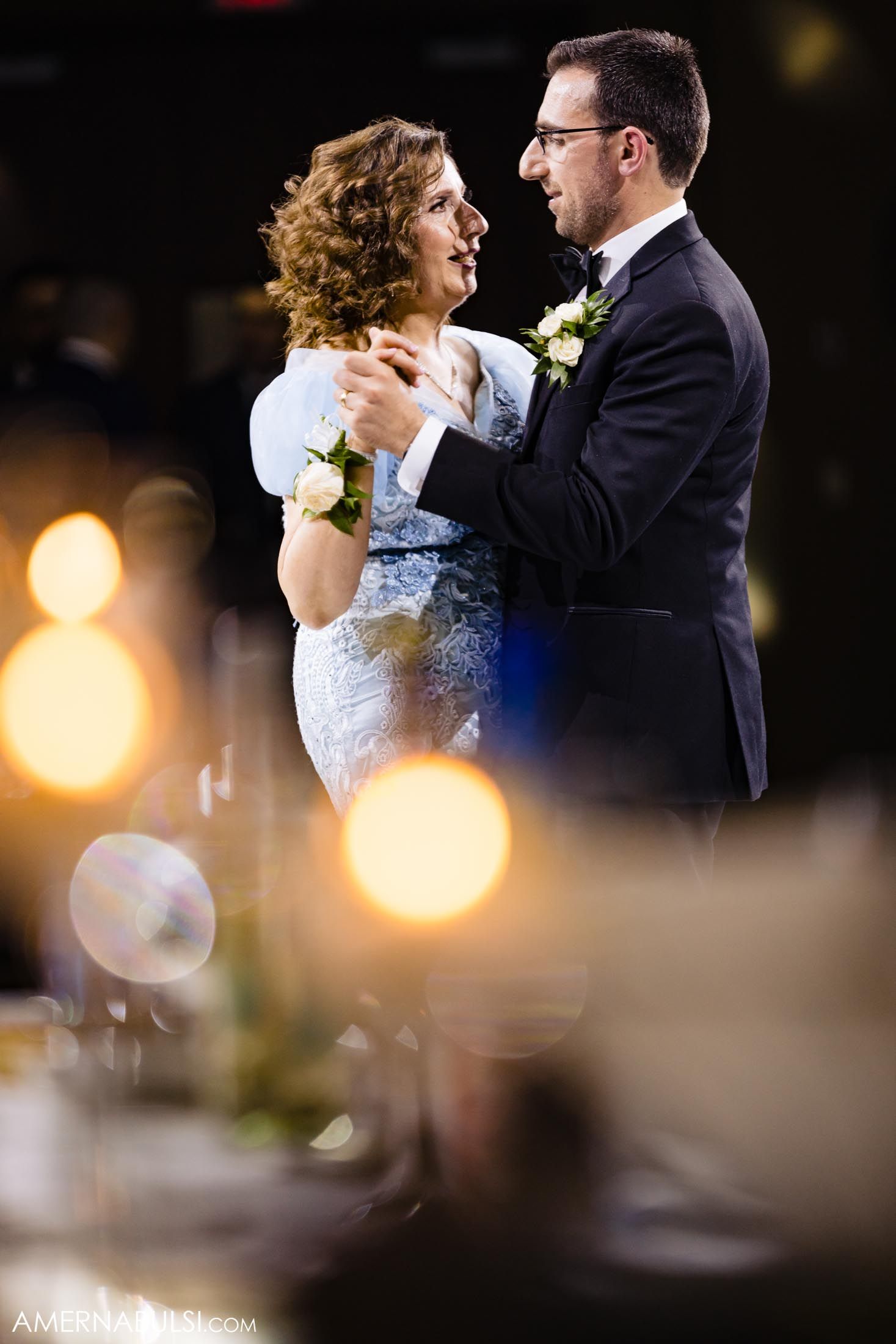 A romantic dance moment captured with soft bokeh lights in the foreground during a wedding reception.