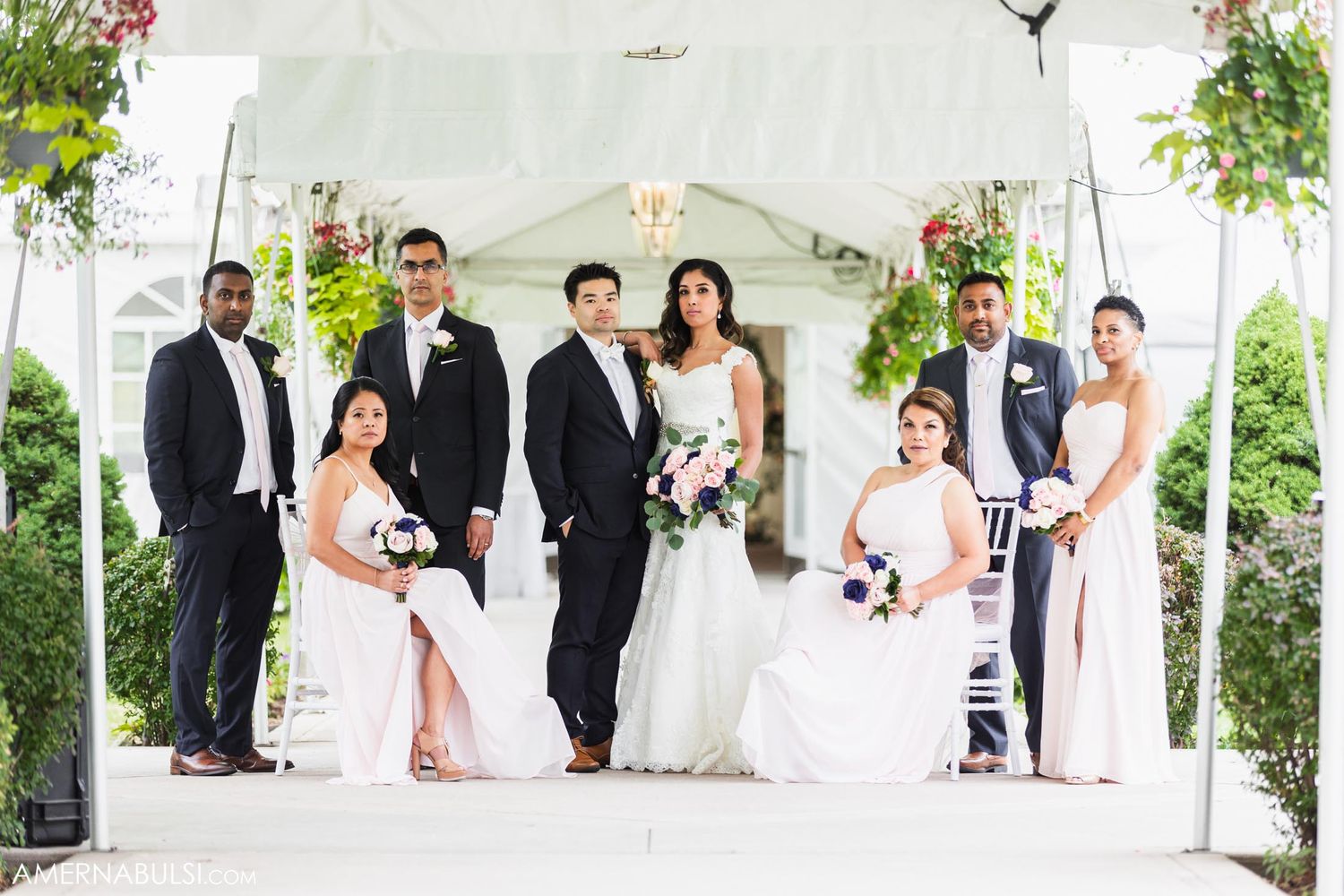 Wedding party poses elegantly under white tent with floral decorations and greenery.