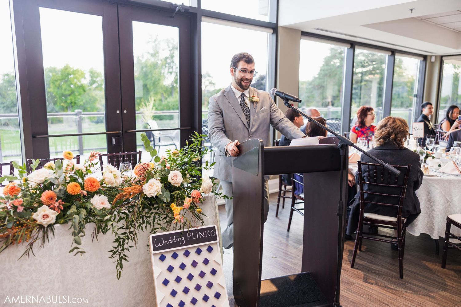 Wedding speaker delivers speech at podium with floral decorations in bright reception venue.