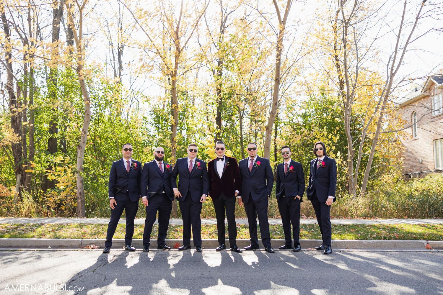Wedding party in formal attire lined up outdoors against autumn trees.