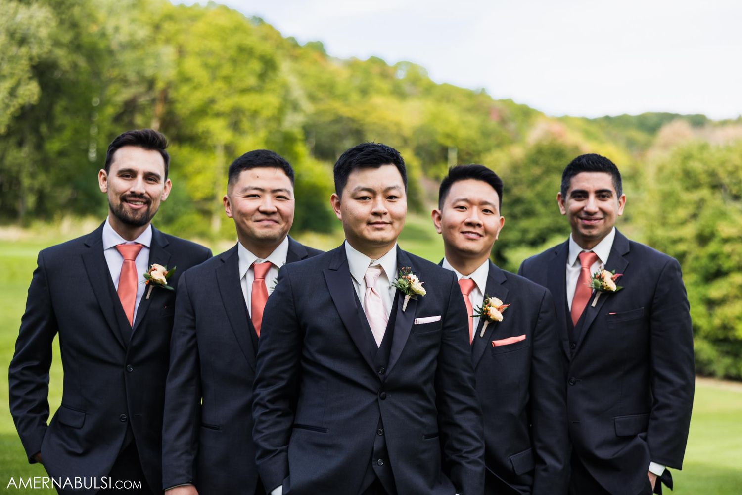 Groomsmen in dark suits with coral ties pose together on golf course.