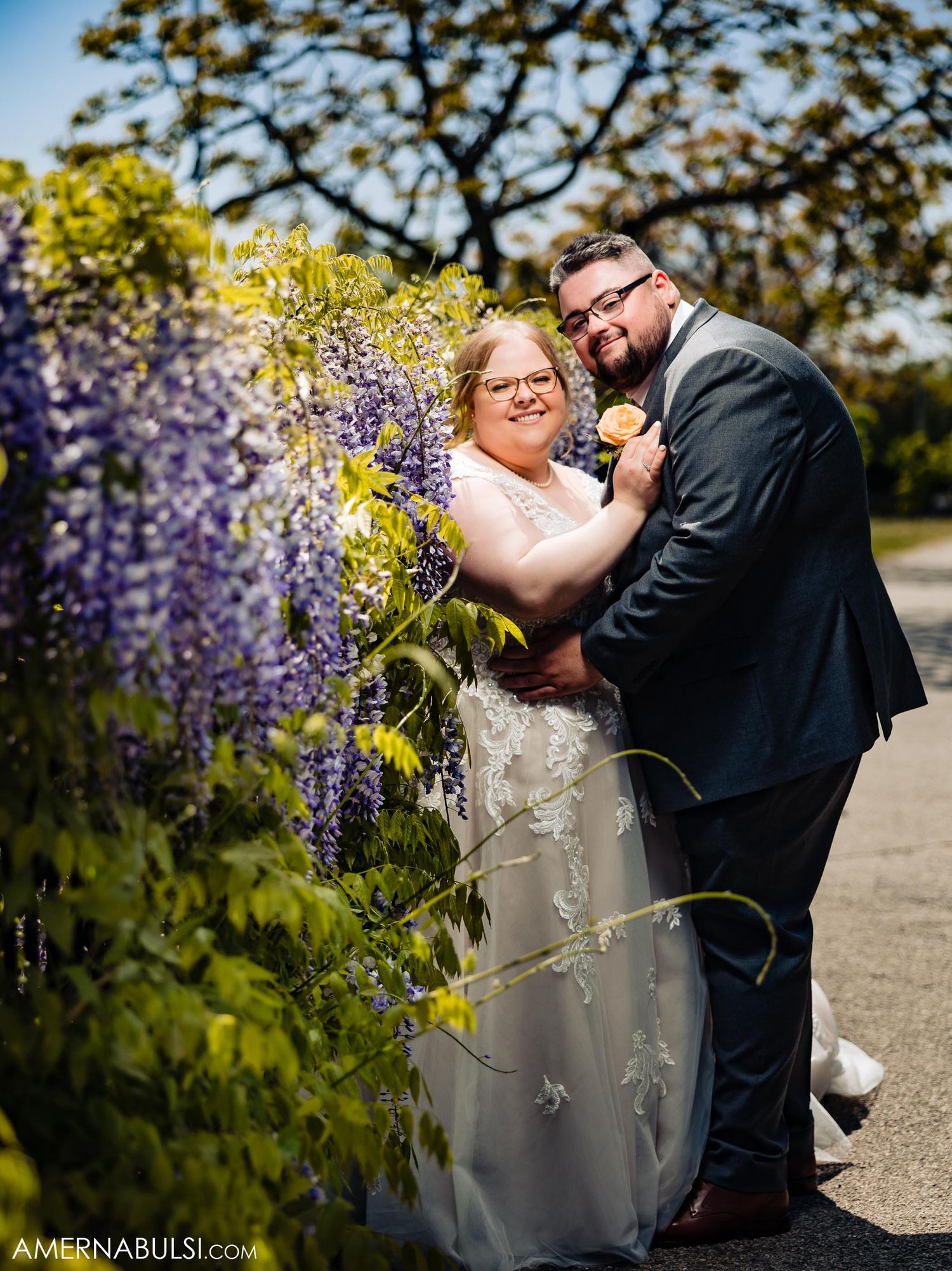 A romantic wedding moment captured beside blooming purple wisteria in a garden setting.