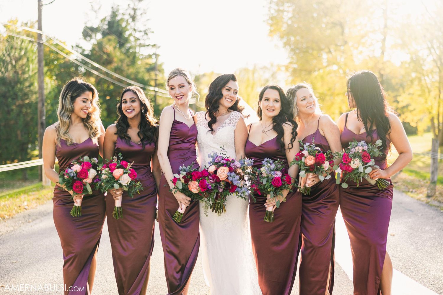 Bridesmaids in burgundy dresses holding pink bouquets in warm sunset lighting.