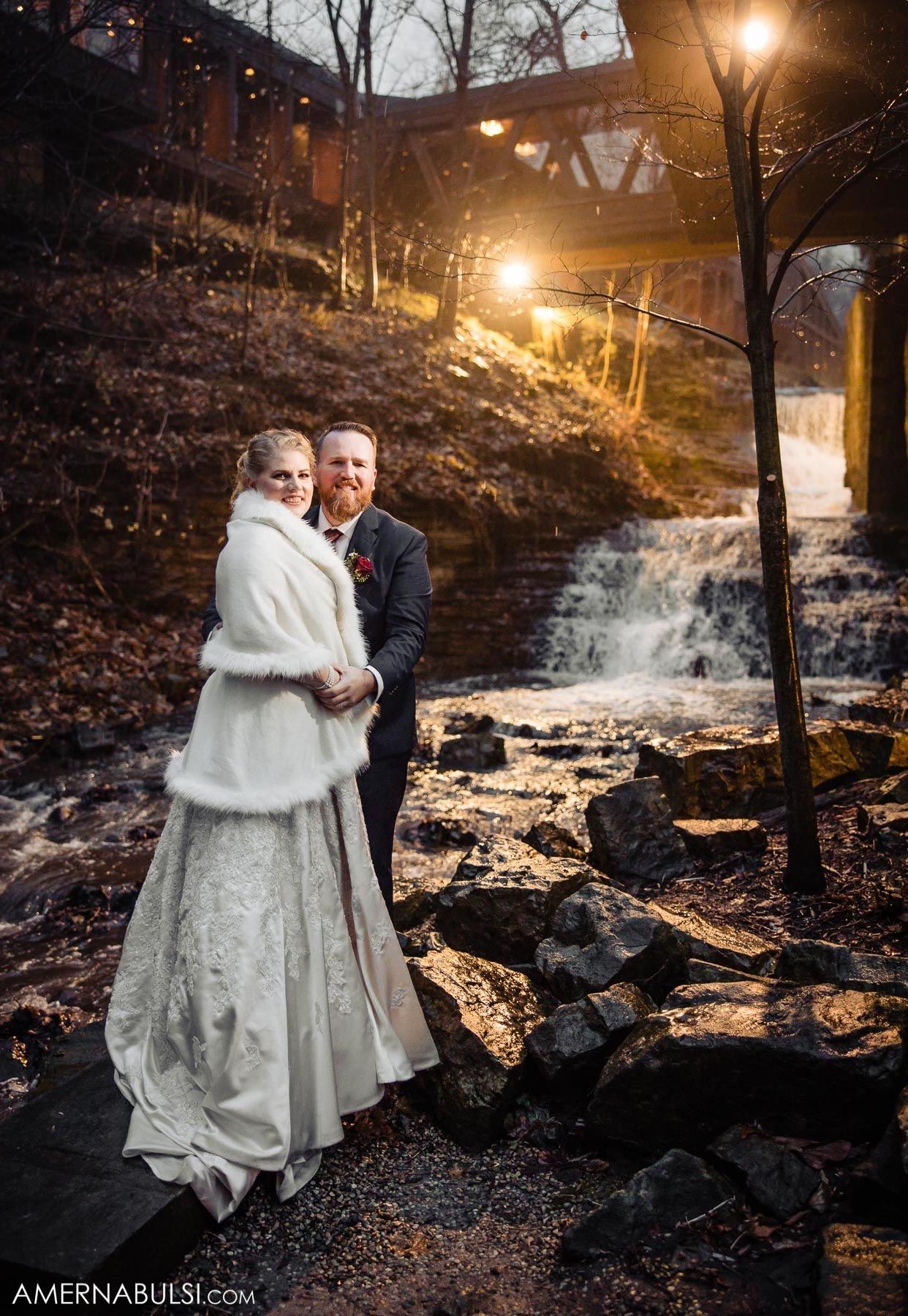Romantic winter wedding photo session at dusk by a waterfall with glowing streetlights creating a magical atmosphere.