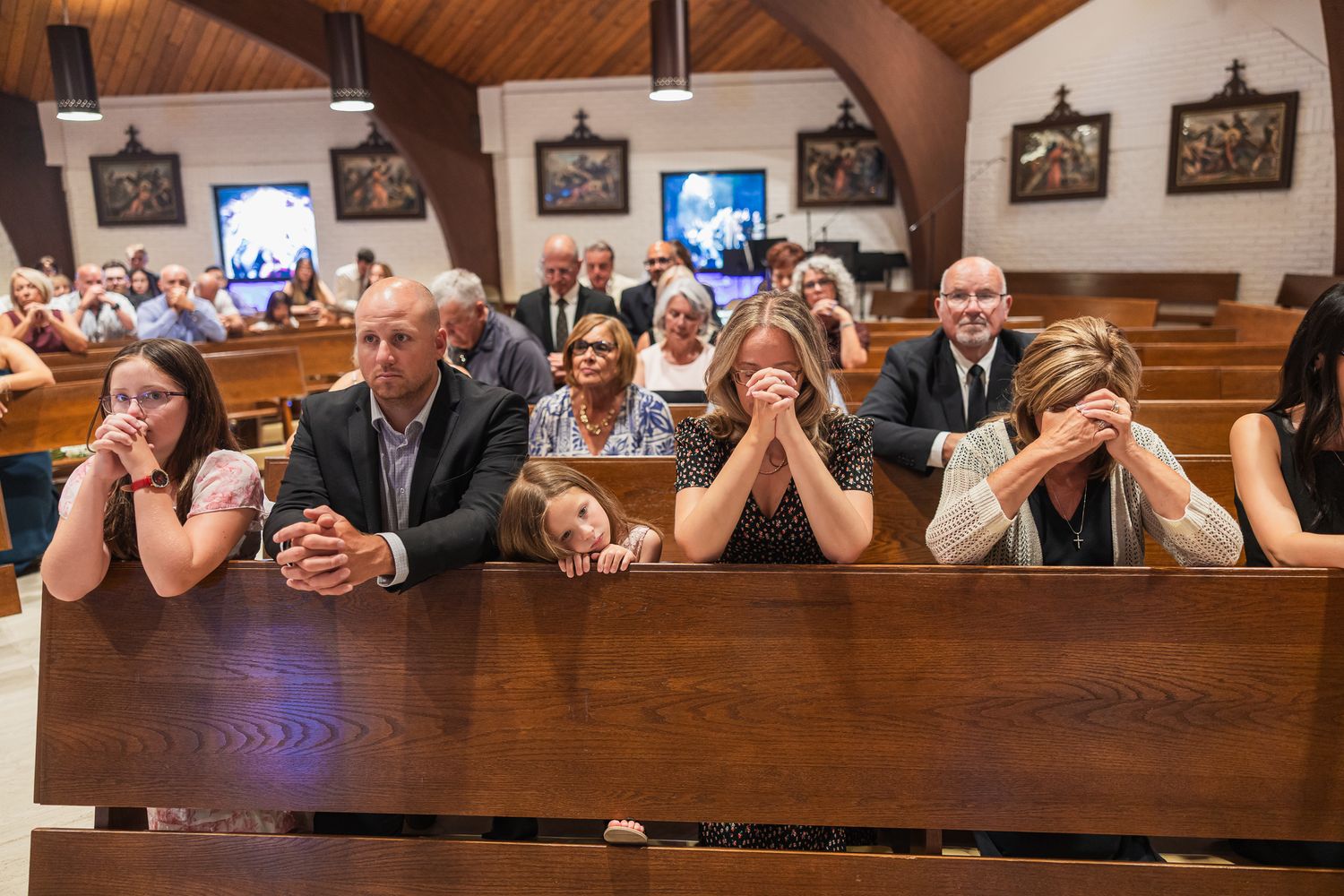Emotional wedding guests seated in church pews, some with heads bowed in prayer during a wedding ceremony.