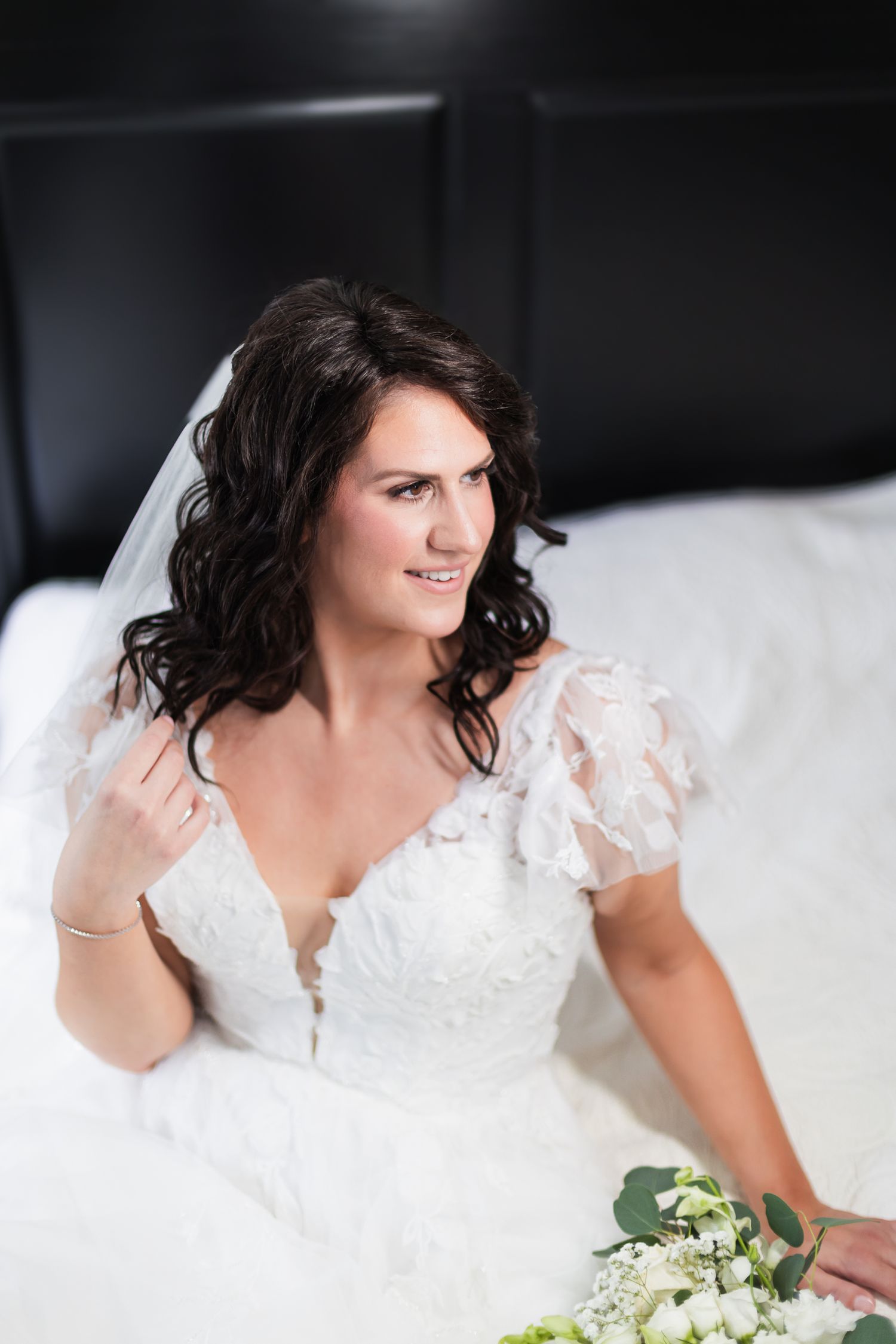 Beautiful bride in lace wedding dress with veil sitting on bed holding white bouquet.