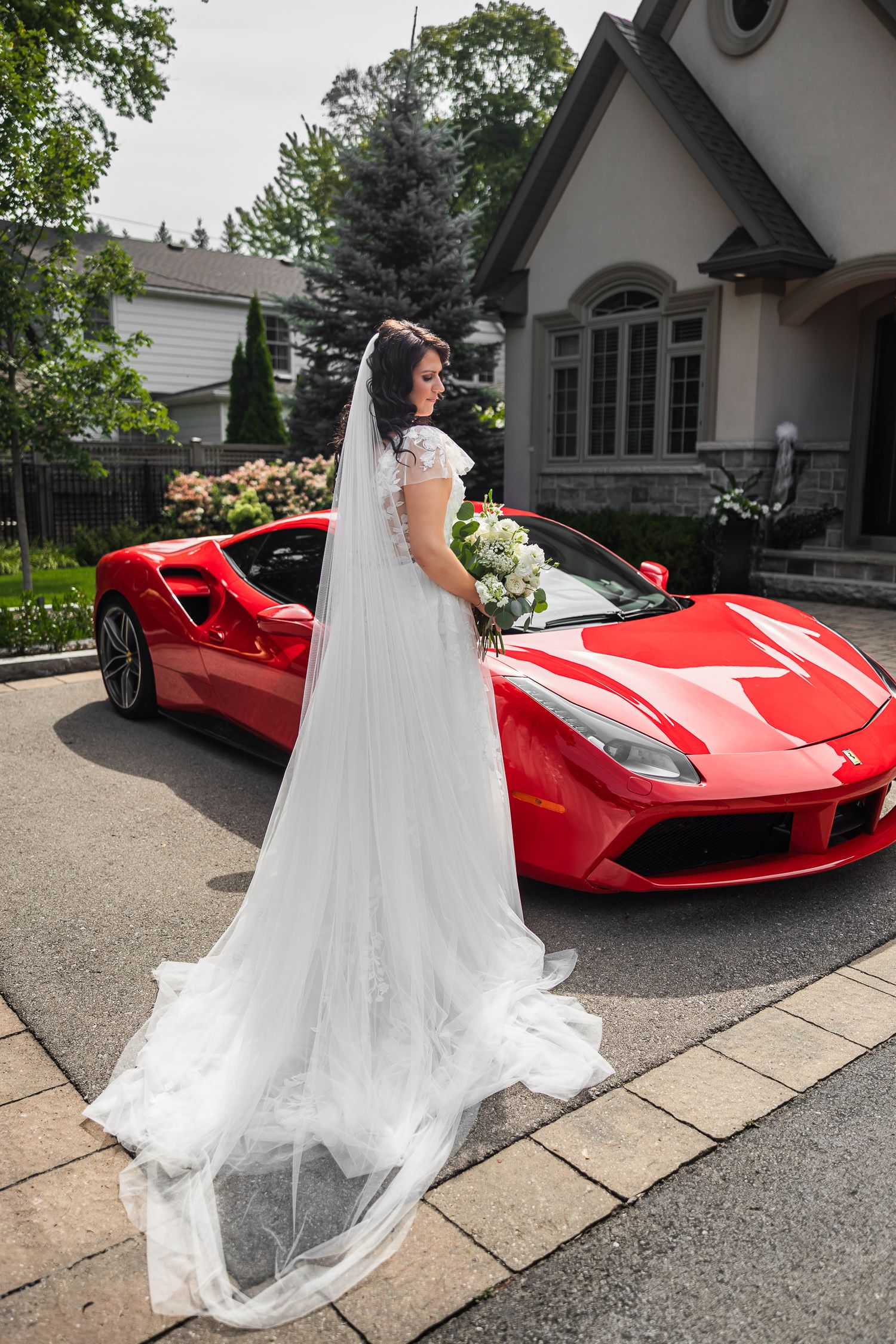Bride in white lace gown with long veil standing beside red Ferrari sports car.