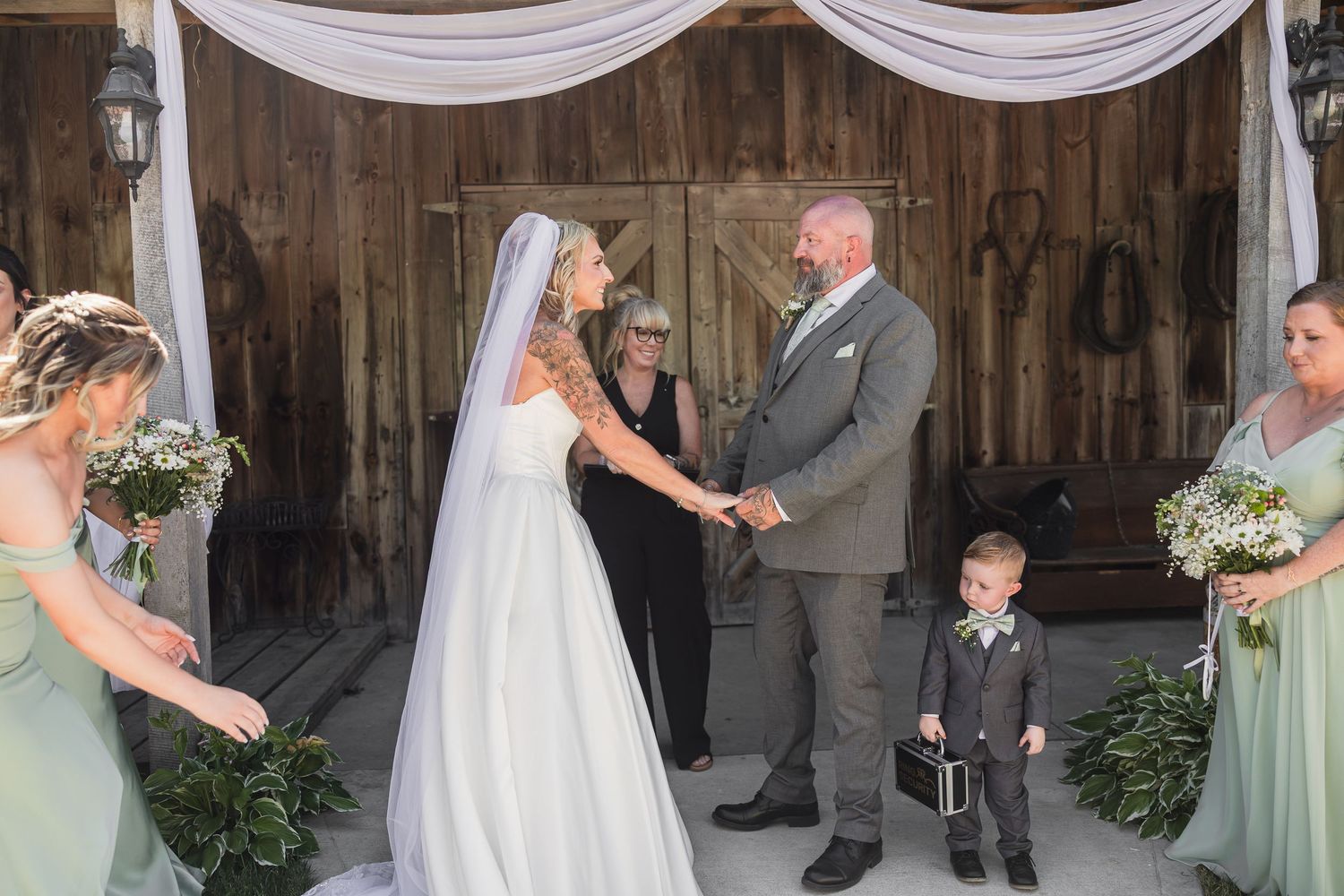 Bride and groom hold hands at rustic barn wedding ceremony with bridesmaids and ring bearer present.