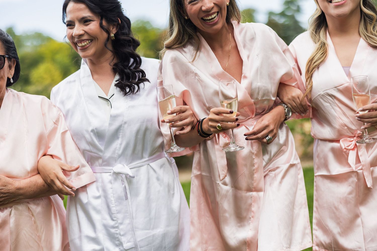 Bridesmaids in pink satin robes laughing and holding champagne glasses outdoors before the wedding.