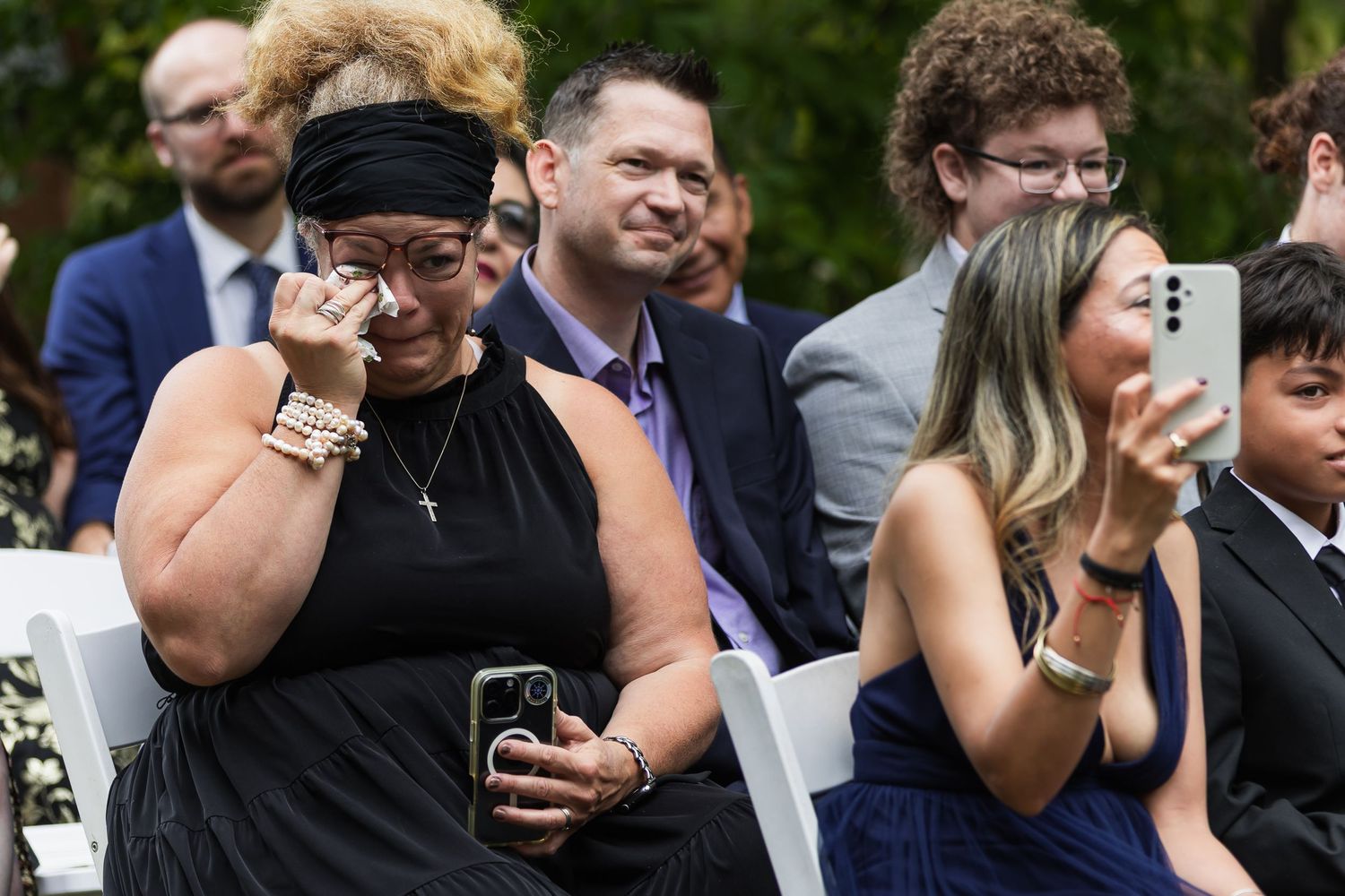 Emotional wedding guest wipes tears while another photographs the outdoor ceremony on a smartphone.