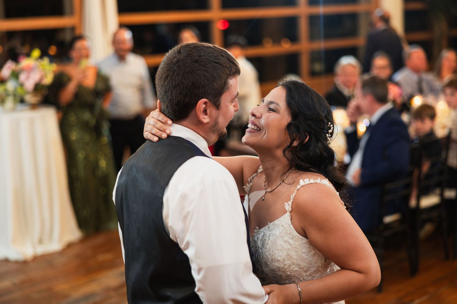 Bride and groom share their first dance, smiling joyfully at their wedding reception with guests applauding in the background.