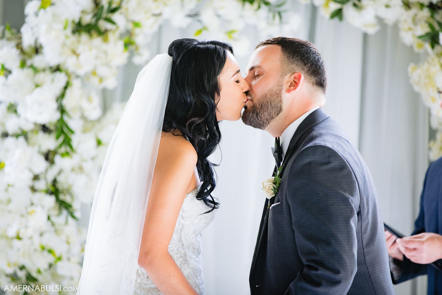 Couple shares their first kiss as newlyweds under an elegant white floral arch.