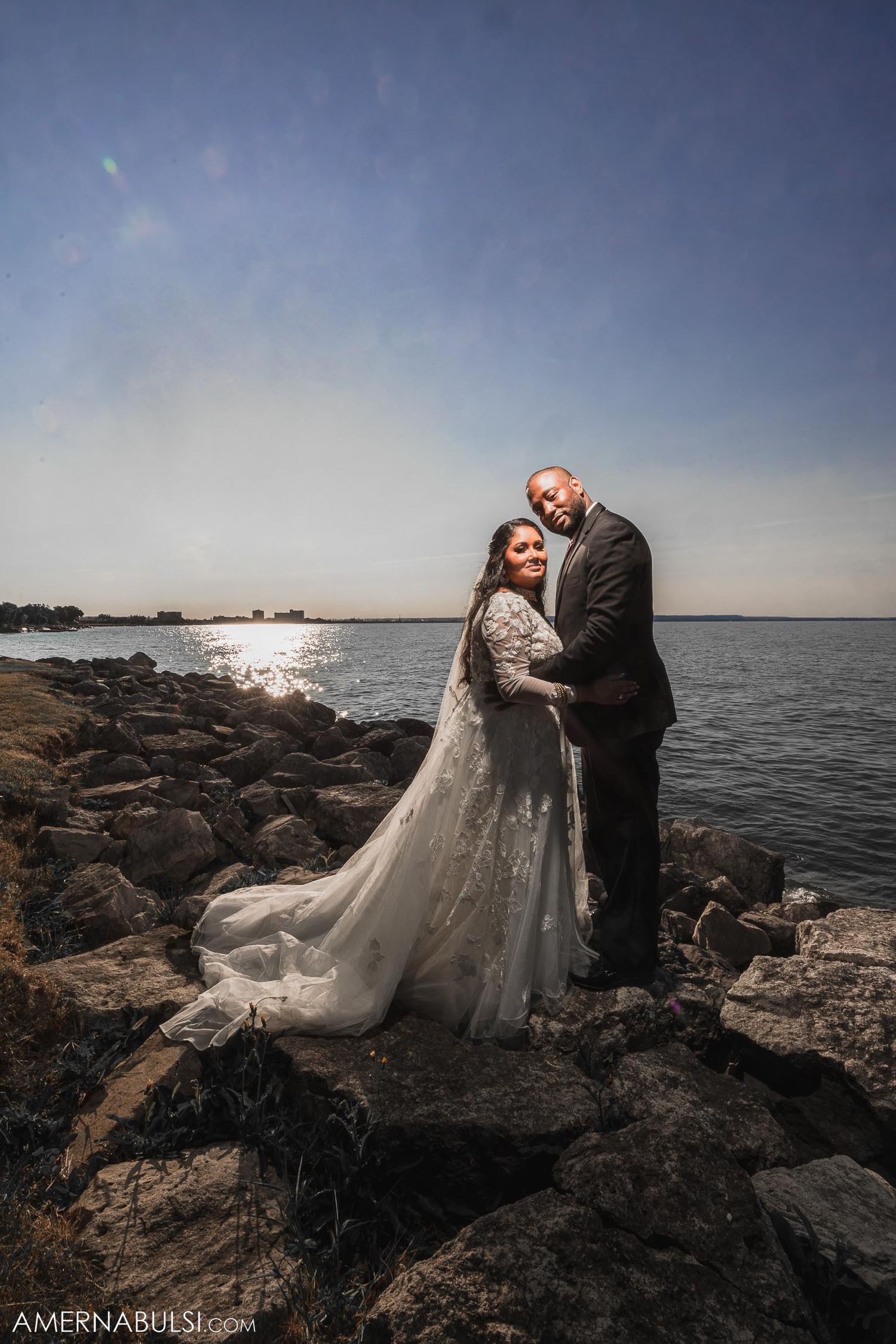 A romantic wedding photo on rocky shoreline with dramatic lighting and water views.